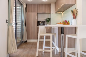 A modern kitchen with wooden cabinets, a white countertop, and a stainless steel sink. There are two white bar stools and a dining table. - Club Del Sole Orbetello Family Collection (Orbetello)
