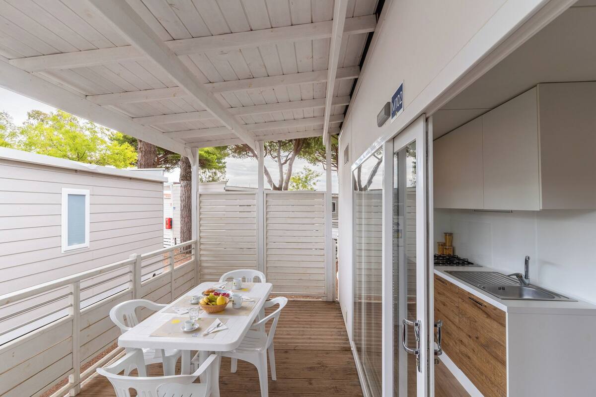 Une terrasse couverte avec une table dressée pour les repas, un coin cuisine équipé de placards et d’un évier, et une vue sur les arbres environnants.