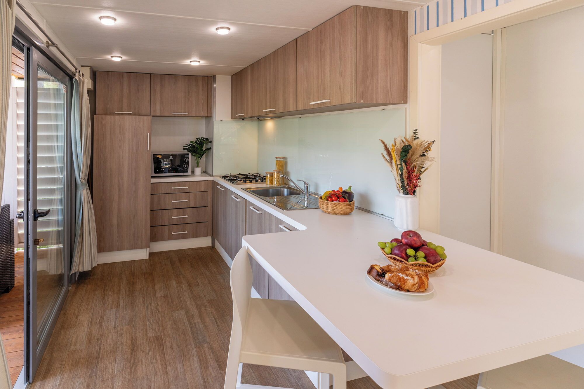 Une cuisine moderne avec des meubles en bois, un four à micro-ondes, un évier et un coin repas comprenant une table et des chaises blanches.