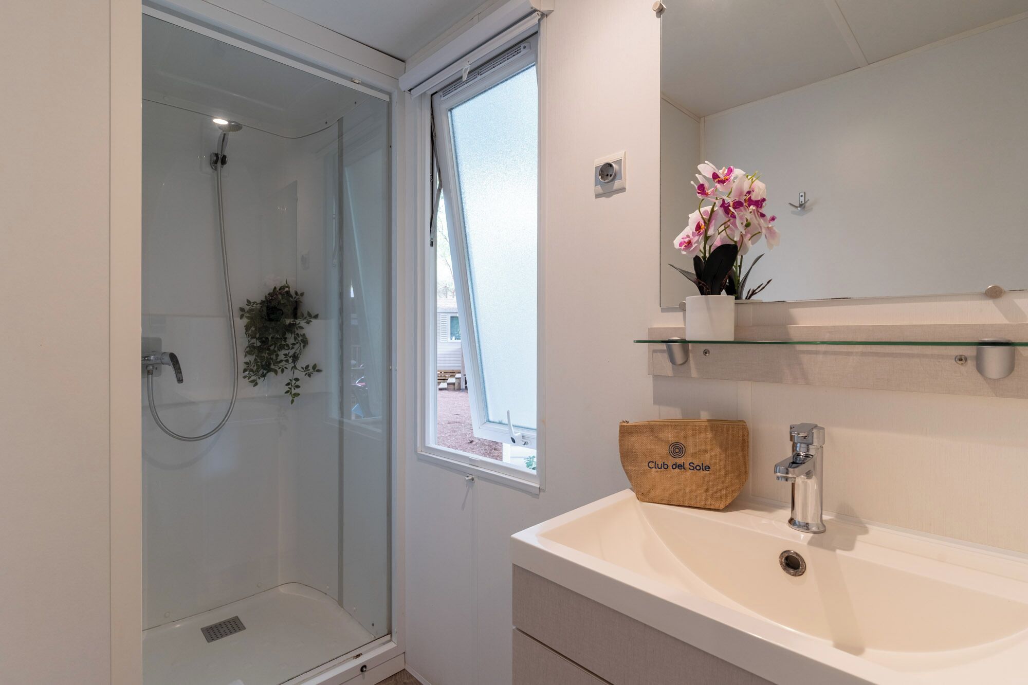 Une salle de bain avec une douche à parois de verre, un lavabo blanc, un miroir et une fenêtre.