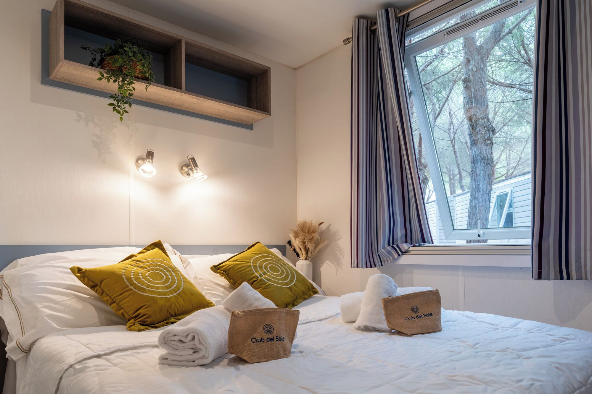Une chambre d’hôtel avec un lit, des oreillers, une serviette, une étagère avec des plantes, des appliques murales et une fenêtre avec des rideaux.