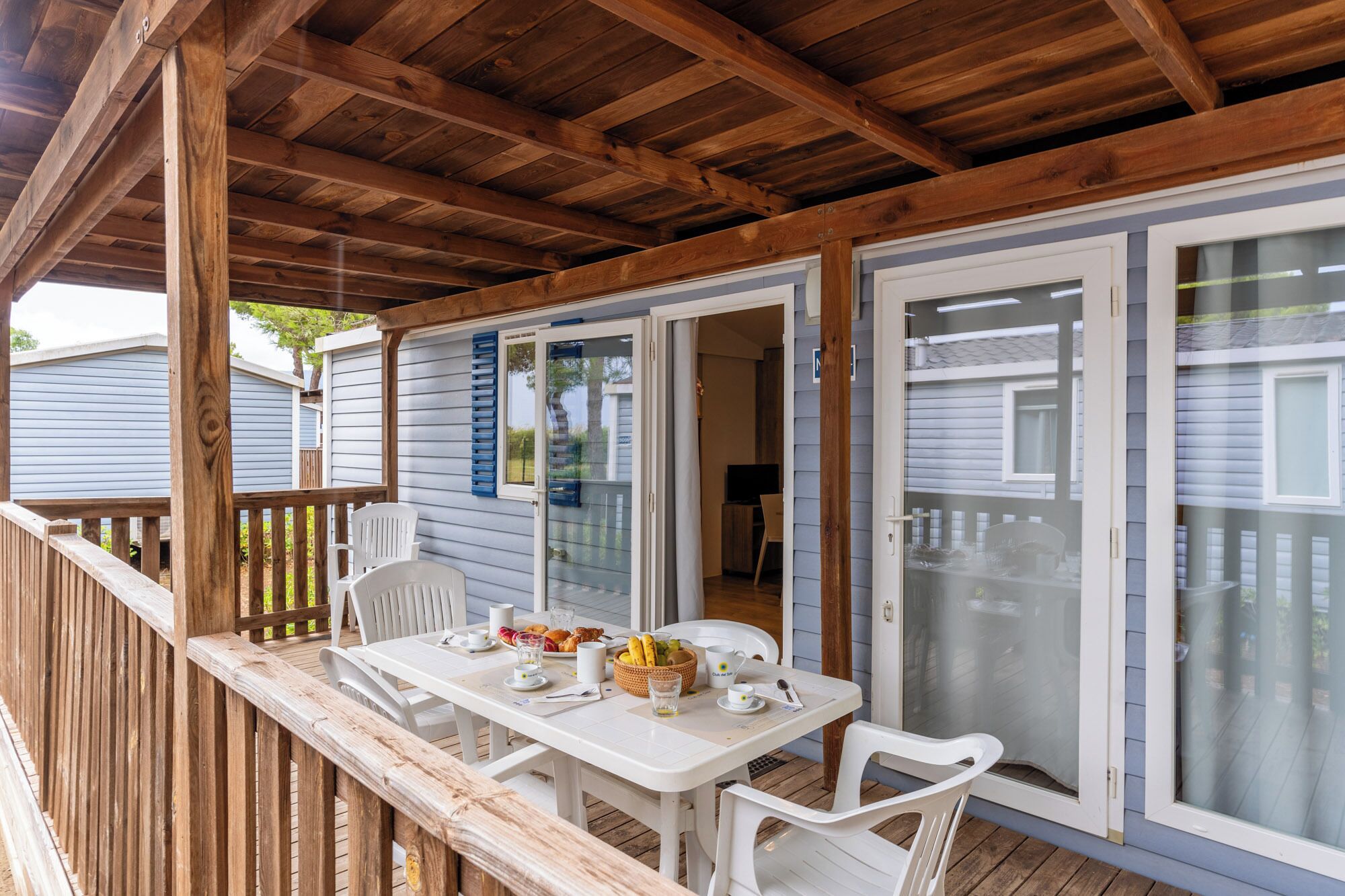 Un espace extérieur abrité, aménagé pour les repas, avec une table et des chaises, une terrasse en bois et une vue sur une maison dotée de portes-fenêtres coulissantes.