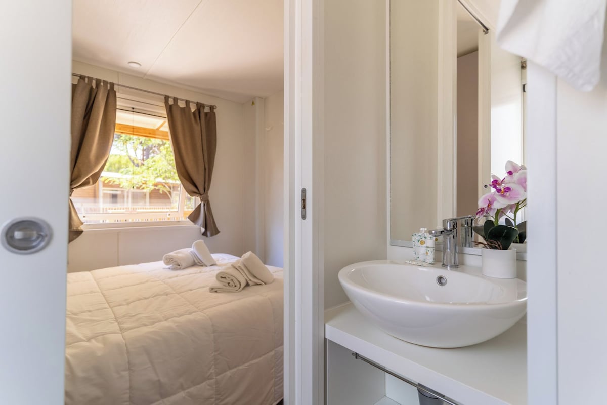 A bedroom with a bed, a window with curtains, and a bathroom with a sink and mirror.