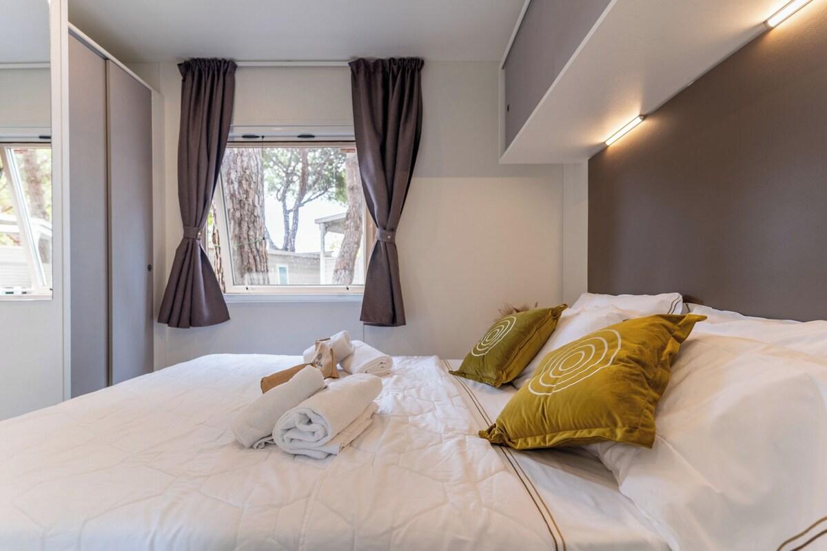 A neatly made bed with pillows and towels, a window with curtains, and a headboard with built-in lighting.