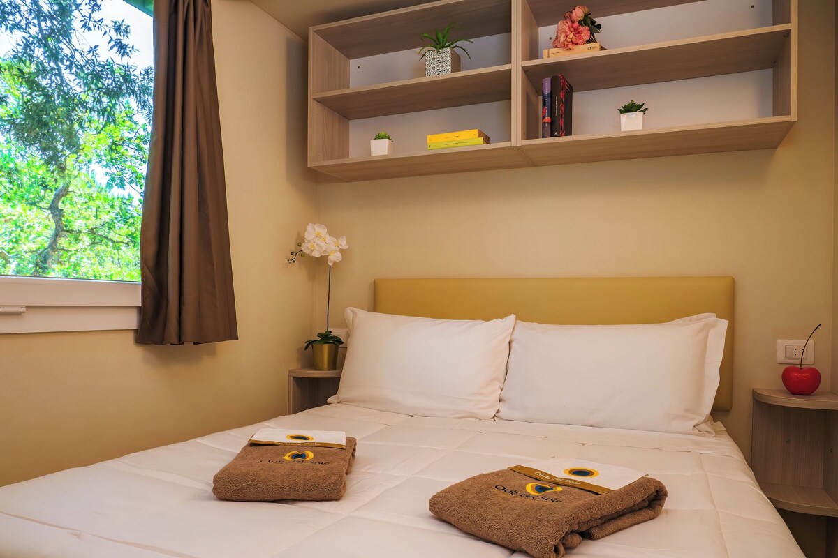 A neatly made bed with two towels, a shelf with books and plants, and a window with curtains.