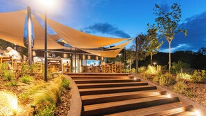 A wooden staircase leading to a covered outdoor seating area with tables and chairs, surrounded by plants and trees.