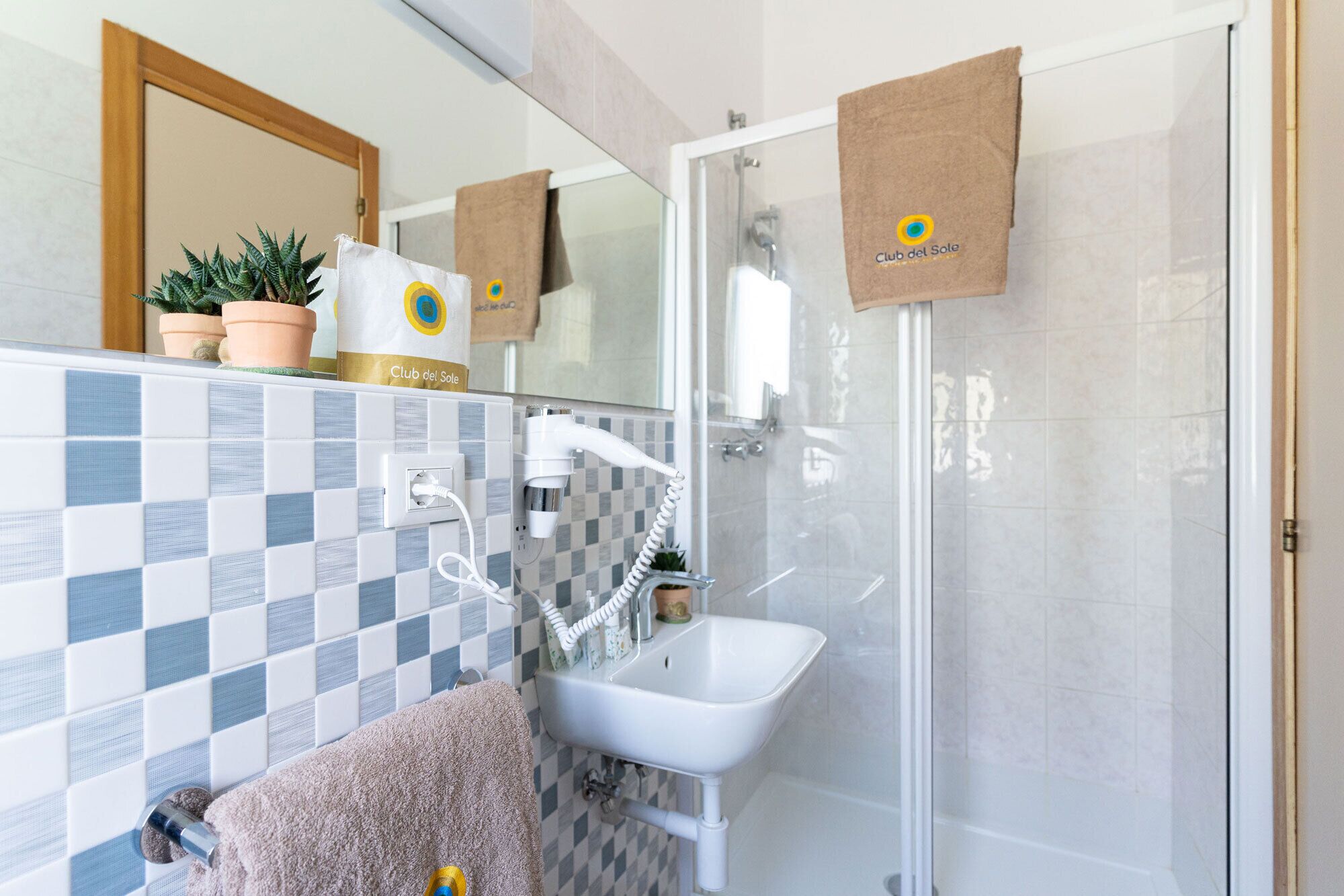 Ein Badezimmer mit Dusche, Waschbecken und Spiegel. Handtücher und ein Topf mit einer Pflanze stehen auf der Ablagefläche.