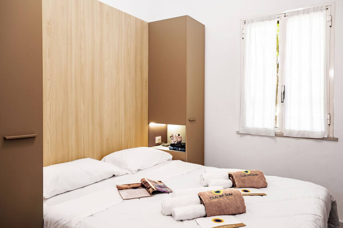 Una camera d'albergo con un letto, lenzuola bianche, una parete rivestita in legno, un comodino con una lampada e una finestra con tende.