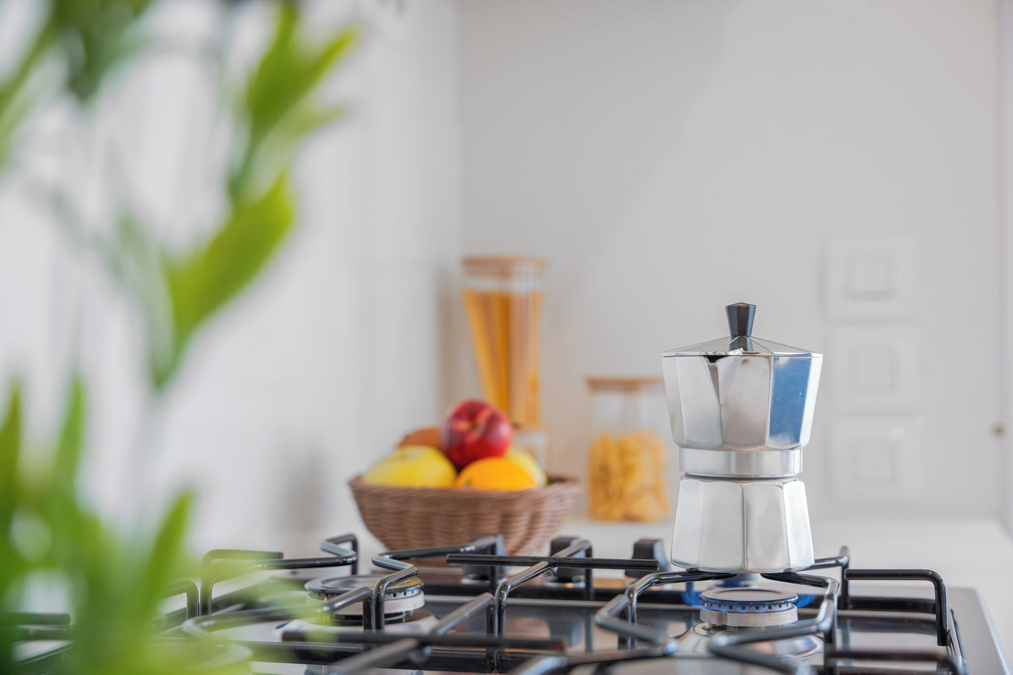 Una cucina con un fornello a gas, una caffettiera e un cesto di frutta sullo sfondo.