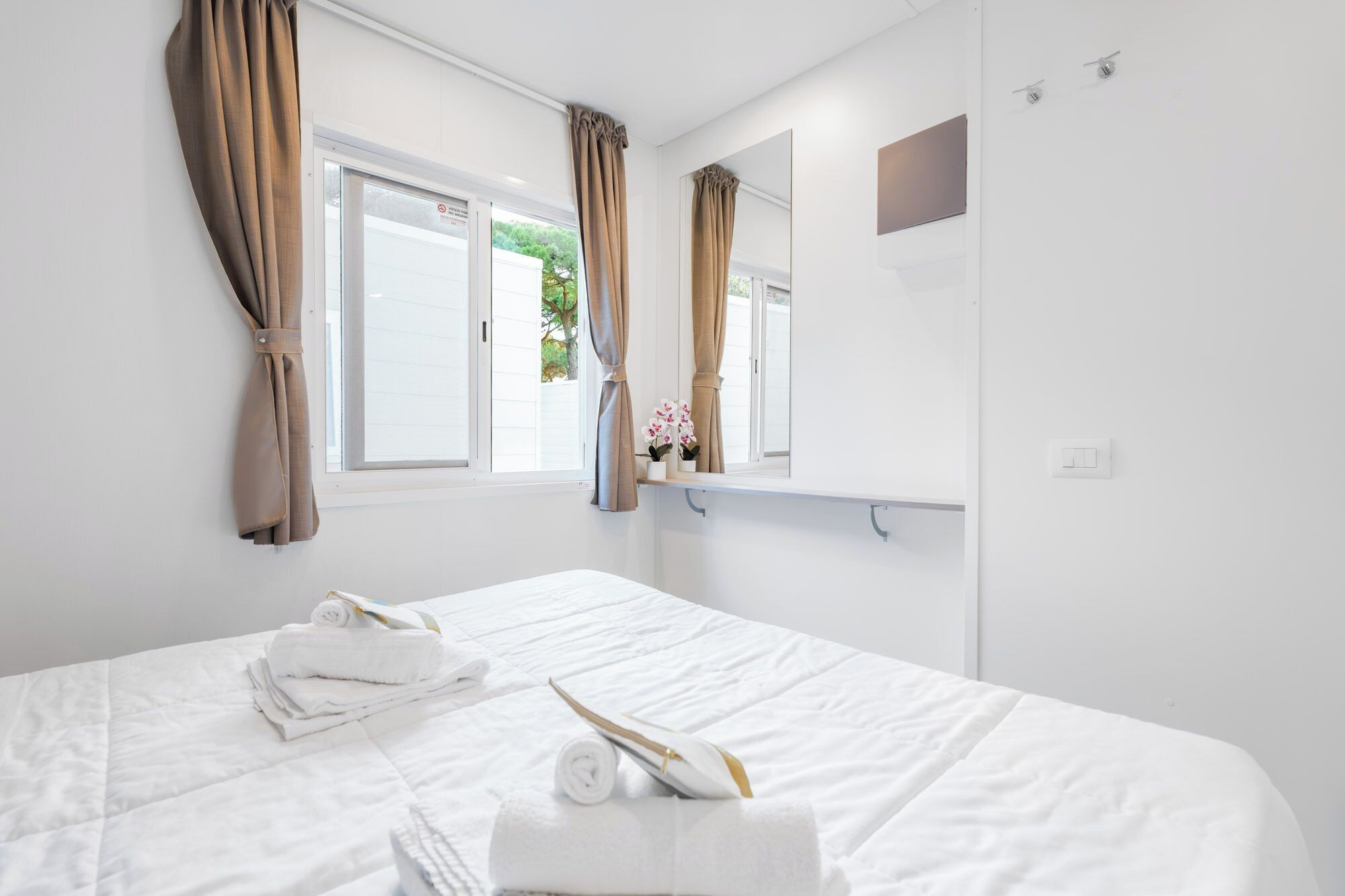 Ein Hotelzimmer mit einem Bett, zwei mit Vorhängen versehenen Fenstern, einem Spiegel und einem kleinen Regal mit einer Pflanze.