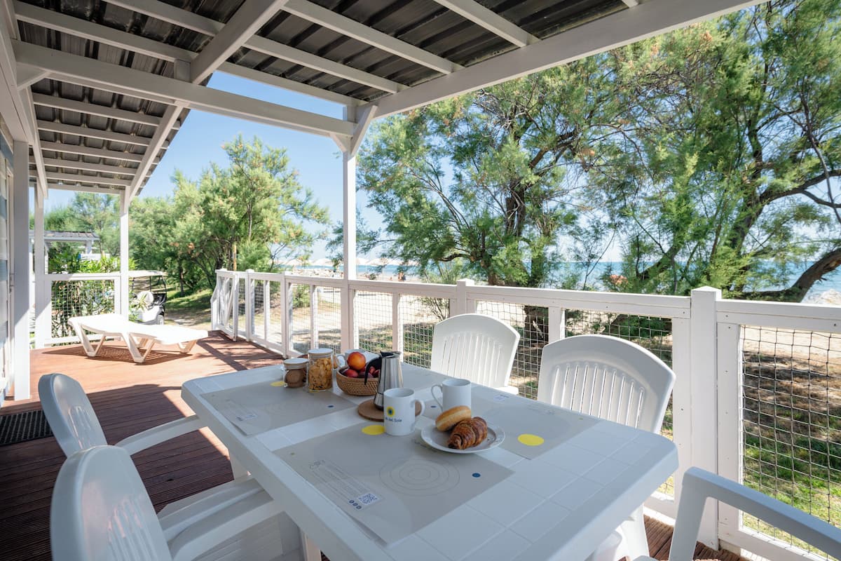 Un'area pranzo all'aperto coperta con mobili bianchi, un tavolo apparecchiato con cibo e bevande e vista sul mare e sugli alberi.