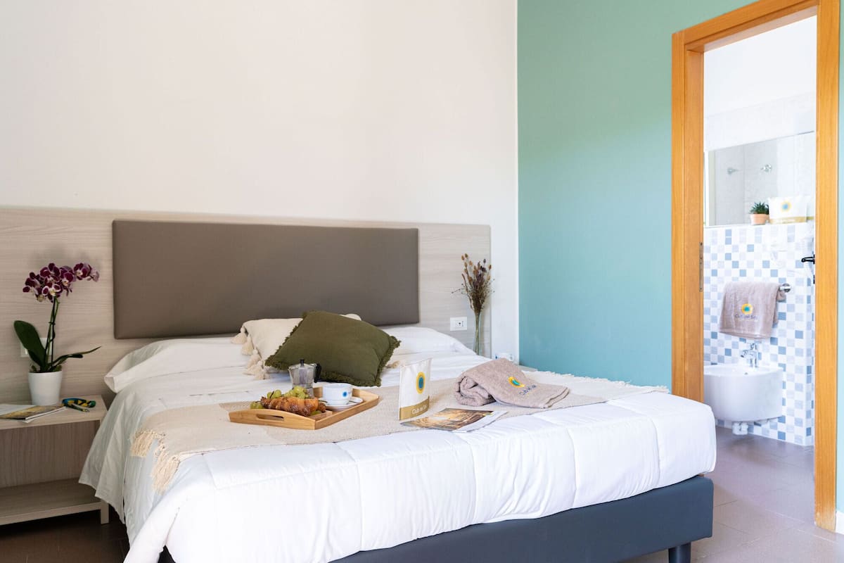 Un letto rifatto con cura, un vassoio con del cibo, un comodino con una pianta e vista su un bagno con parete in piastrelle a scacchi.
