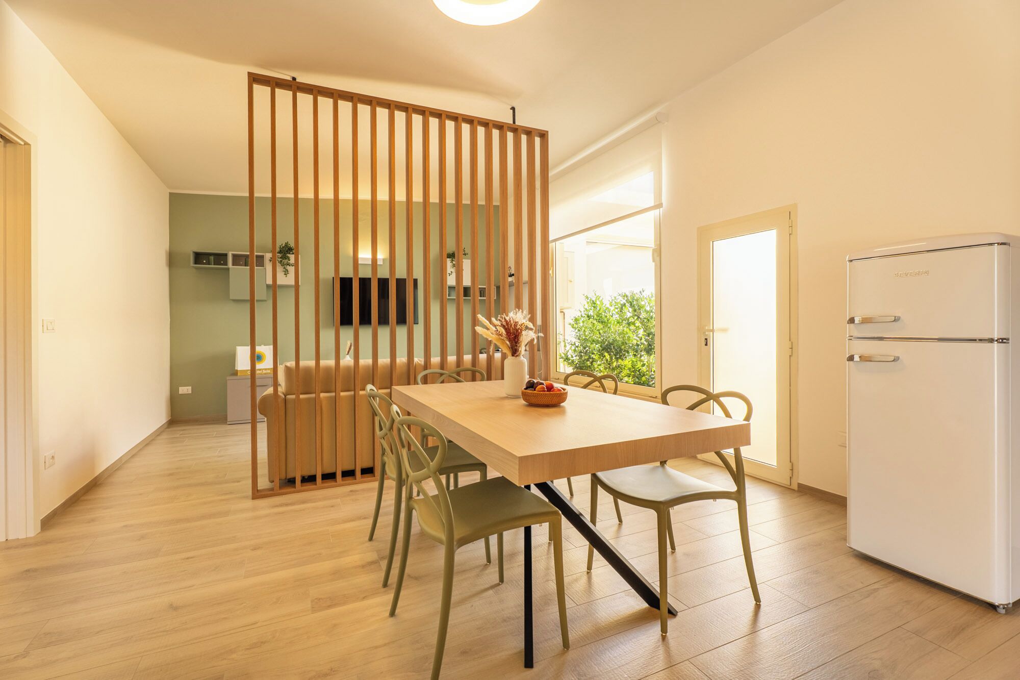 Eine moderne Küche mit Essbereich, Holzlamellen, einem weißen Kühlschrank und Blick auf Grünfläche.