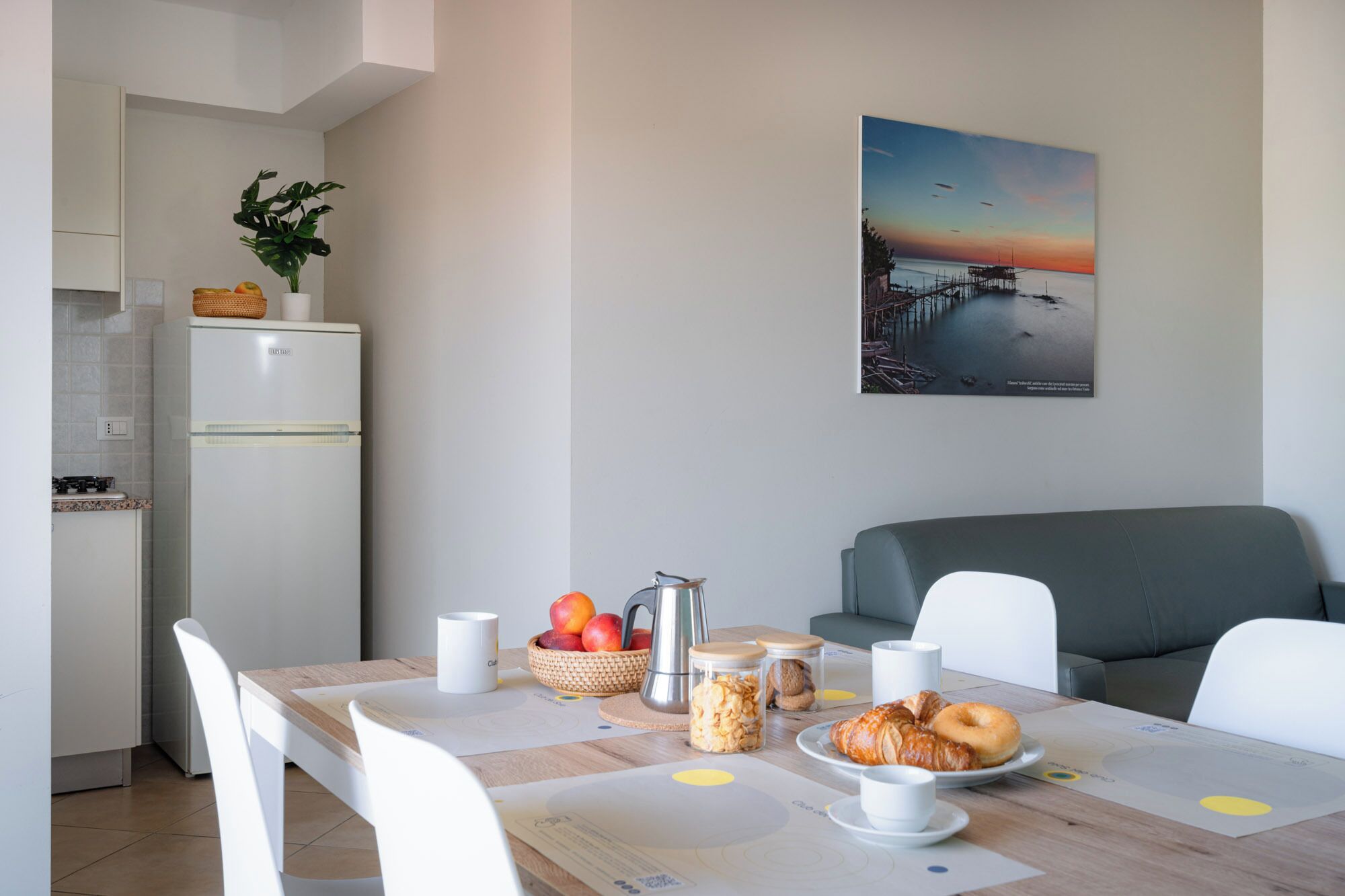 Un'area pranzo con tavolo apparecchiato per la colazione, una cucina sullo sfondo e un dipinto del tramonto sulla parete.