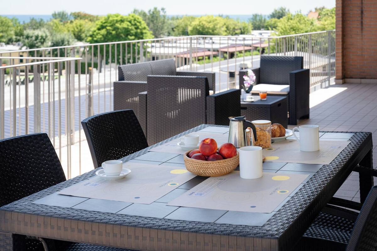 Tavola imbandita per la colazione, con frutta in un cesto, una caffettiera e un cornetto, su un patio esterno con mobili in vimini e vista sul mare.