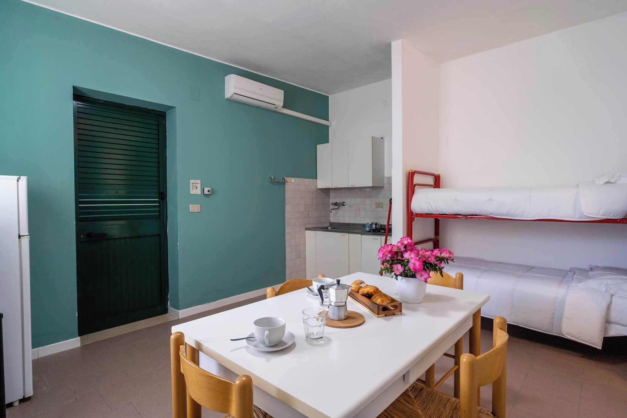Ein kleines Zimmer mit Küchenzeile, einem Esstisch mit Stühlen, einem Stockbett und einem kleinen Tisch mit Blumenvase.