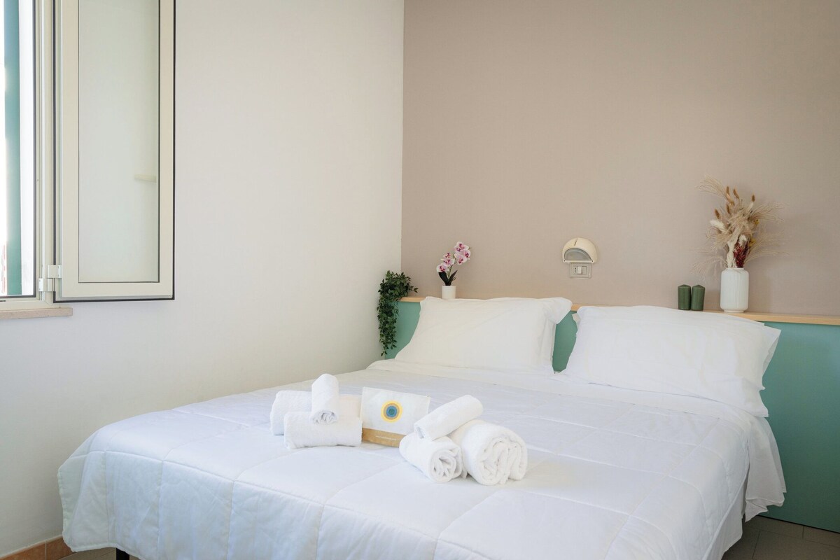 Una cama bien hecha con ropa blanca y toallas, una planta en la mesita de noche y una lámpara de pared.