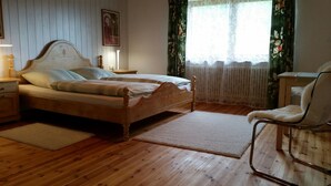 A bedroom with a wooden bed, a nightstand, a chair, and a window with floral curtains. - Tannehüsle large - quiet, with a south-facing view (St. Märgen)