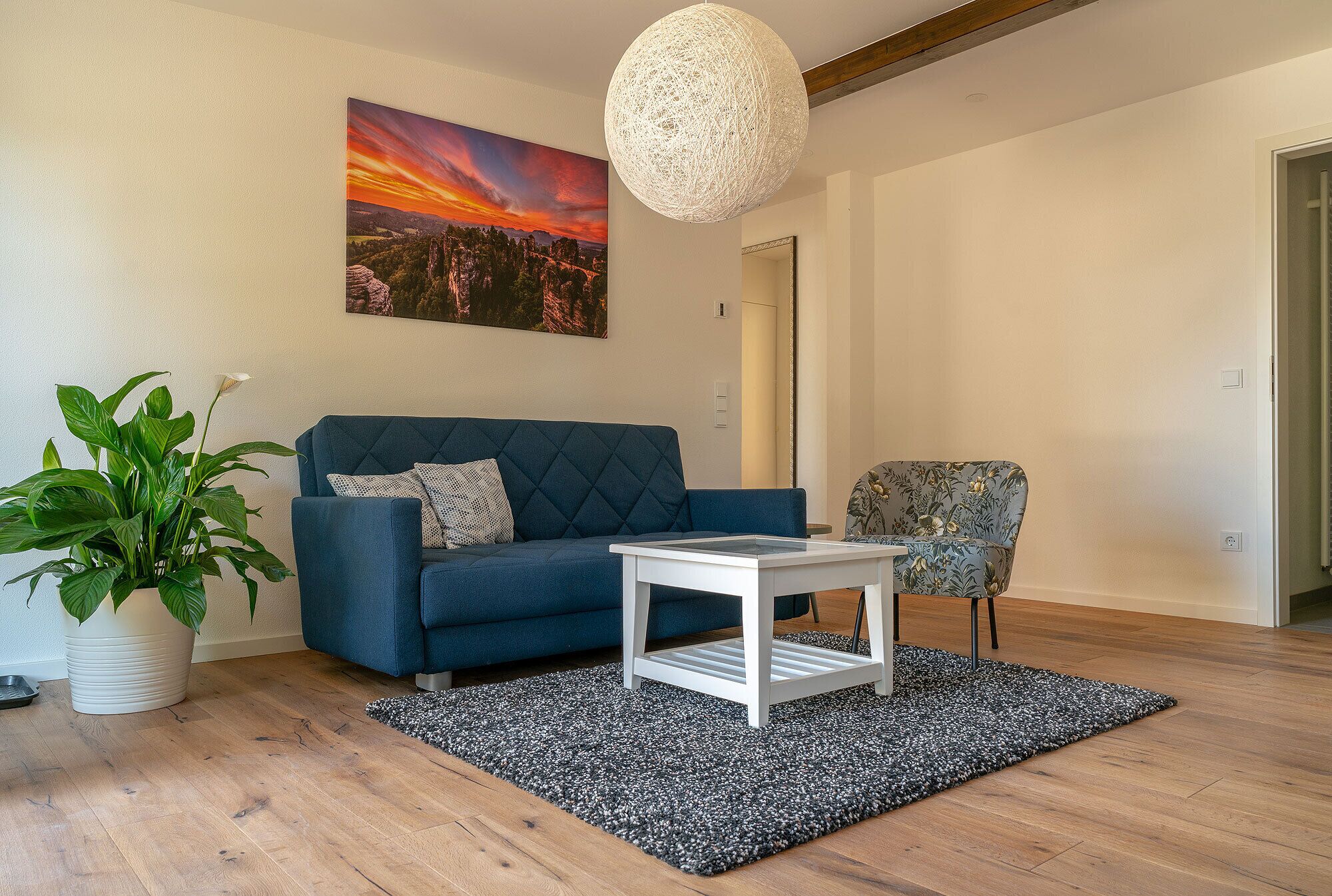 A modern living room with a blue sofa, a white coffee table, a patterned armchair, a large potted plant, and a painting of a sunset.