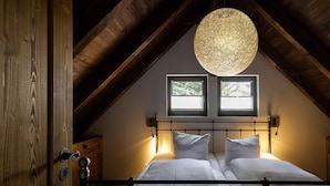 Un dormitorio con techo inclinado, una cama con ropa blanca, dos ventanas y una lámpara colgante.