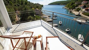 A balcony with two wooden chairs and a table, overlooking a marina with boats and a coastal landscape.