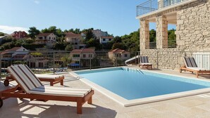 A swimming pool with lounge chairs, a stone building with a balcony, and houses in the background.