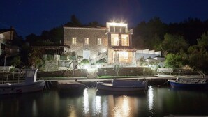 A stone building with a balcony and a lit upper floor, situated by a calm body of water with boats docked in the foreground.