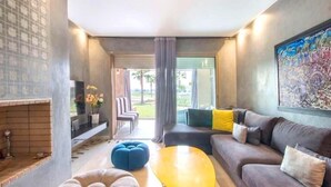 Ein modernes Wohnzimmer mit Sofa, Hocker und Couchtisch. Ein Kamin und ein Blick ins Freie durch große Fenster sind zu sehen.