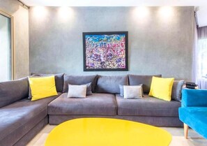 A modern living room with a grey sectional sofa, yellow and grey cushions, a colorful abstract painting on the wall, and a bright yellow coffee table. - Splendid apartment in Prestigia Corail 4675 (Marrakech)