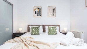 Una cama bien hecha con una manta color beige, dos almohadas con estampado de hojas y una toalla enrollada. Hay dos mesitas de noche con lámparas y dos obras de arte enmarcadas en la pared.