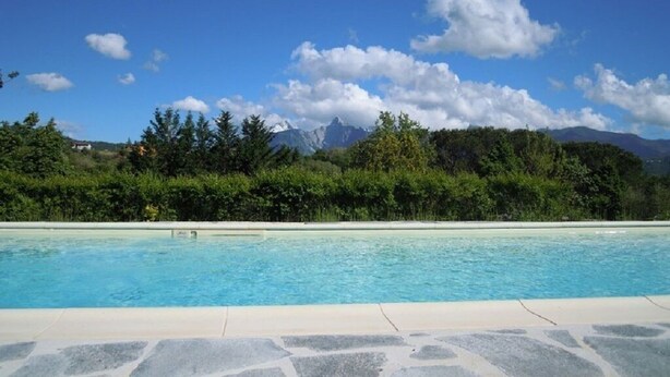 Uma piscina com água azul cristalina, ladeada por um deck de pedra. Ao fundo, uma paisagem verdejante com árvores e uma cordilheira sob um céu azul com nuvens.