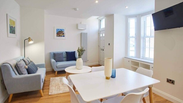 Un salón moderno con un sofá blanco, un sofá azul, una mesa de comedor con sillas, una lámpara de pie y un televisor montado en la pared.