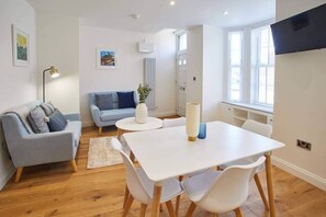 Ein modernes Wohnzimmer mit einem weißen Sofa, einem blauen Sofa, einem Esstisch mit Stühlen, einer Stehlampe und einem an der Wand befestigten Fernseher.