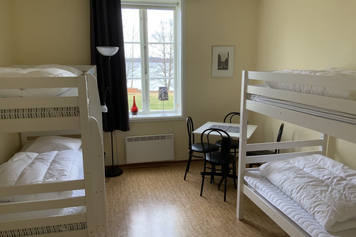 Huoneessa on kerrossängyt, pöytä, tuolit, lamppu, ikkuna, josta on näkymä, ja patteri.