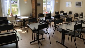 Un salón de conferencias con mesas y sillas, ventanas amplias con cortinas y lámparas fijadas a la pared.