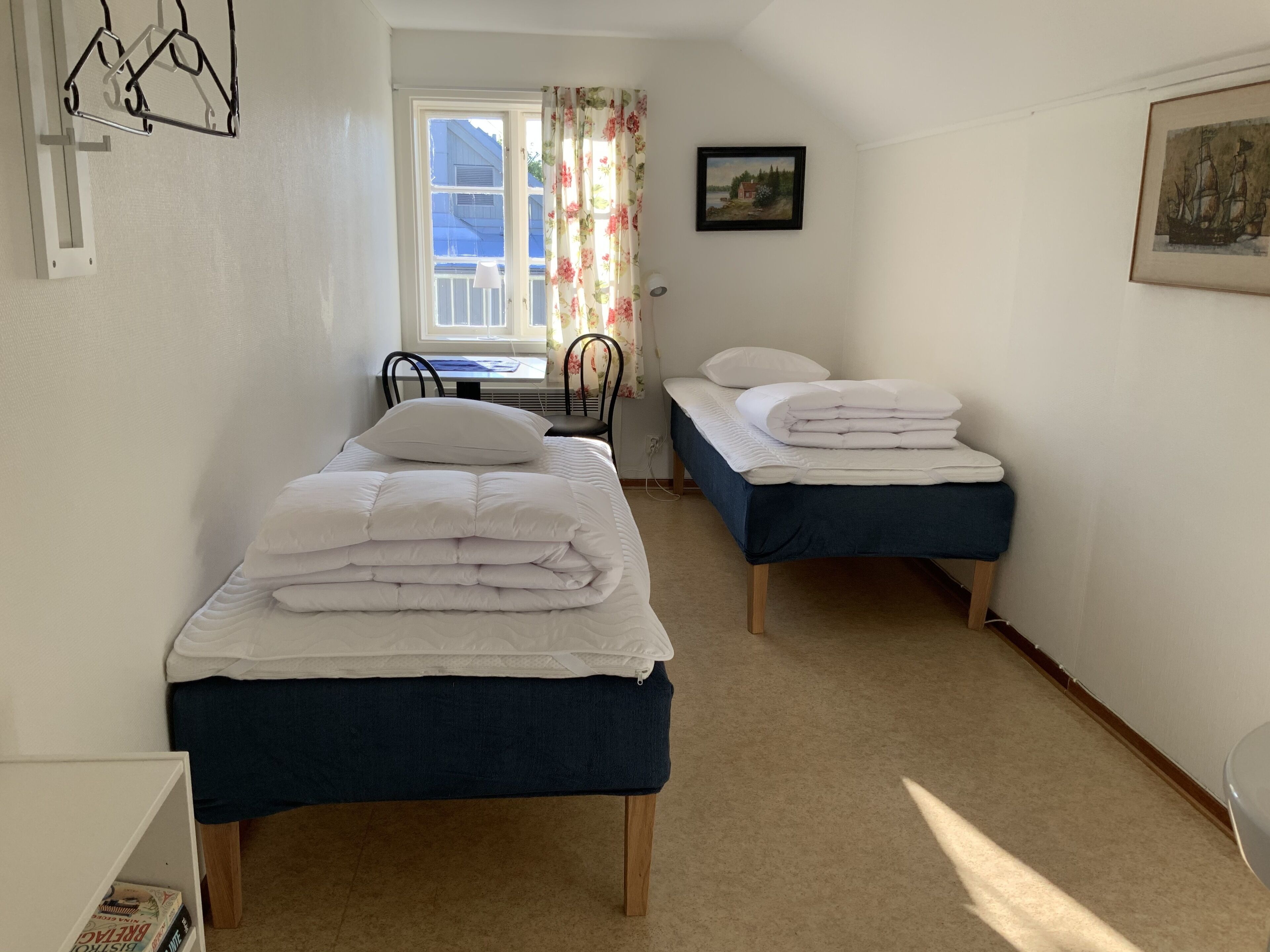 A small, well-lit room with two single beds, a desk with a chair, a window with curtains, and a framed picture on the wall.