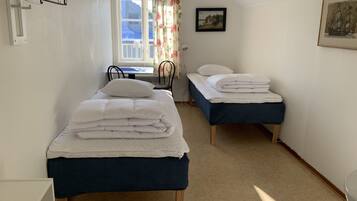 A small, well-lit room with two single beds, a desk with a chair, a window with curtains, and a framed picture on the wall.