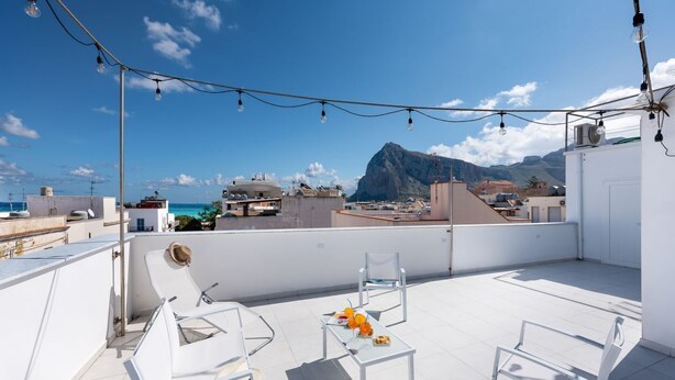 Una terraza en la azotea con sillas blancas, una mesa con frutas y luces colgantes.