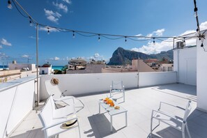 Una terraza en la azotea con sillas blancas, una mesa con frutas y luces colgantes.