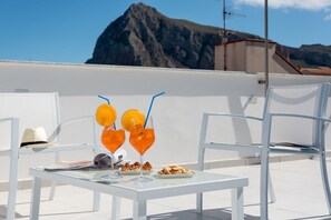 Una mesa con bebidas y aperitivos en una terraza en la azotea, con sillas blancas y una vista a la montaña de fondo.