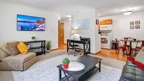 A modern living room with a sofa, coffee table, and TV. There is a kitchen area with a dining table and chairs.