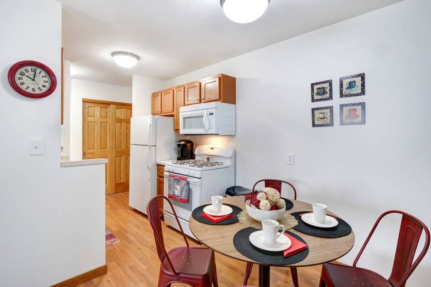 A compact kitchen with a microwave, stove, and refrigerator. There is a dining table with four chairs and a clock on the wall.