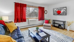 Ein Wohnzimmer mit einem blauen Sofa, roten Vorhängen, einem kleinen Tisch mit einer Pflanze, einem gerahmten Bild an der Wand und einer schwarzen TV-Konsole.