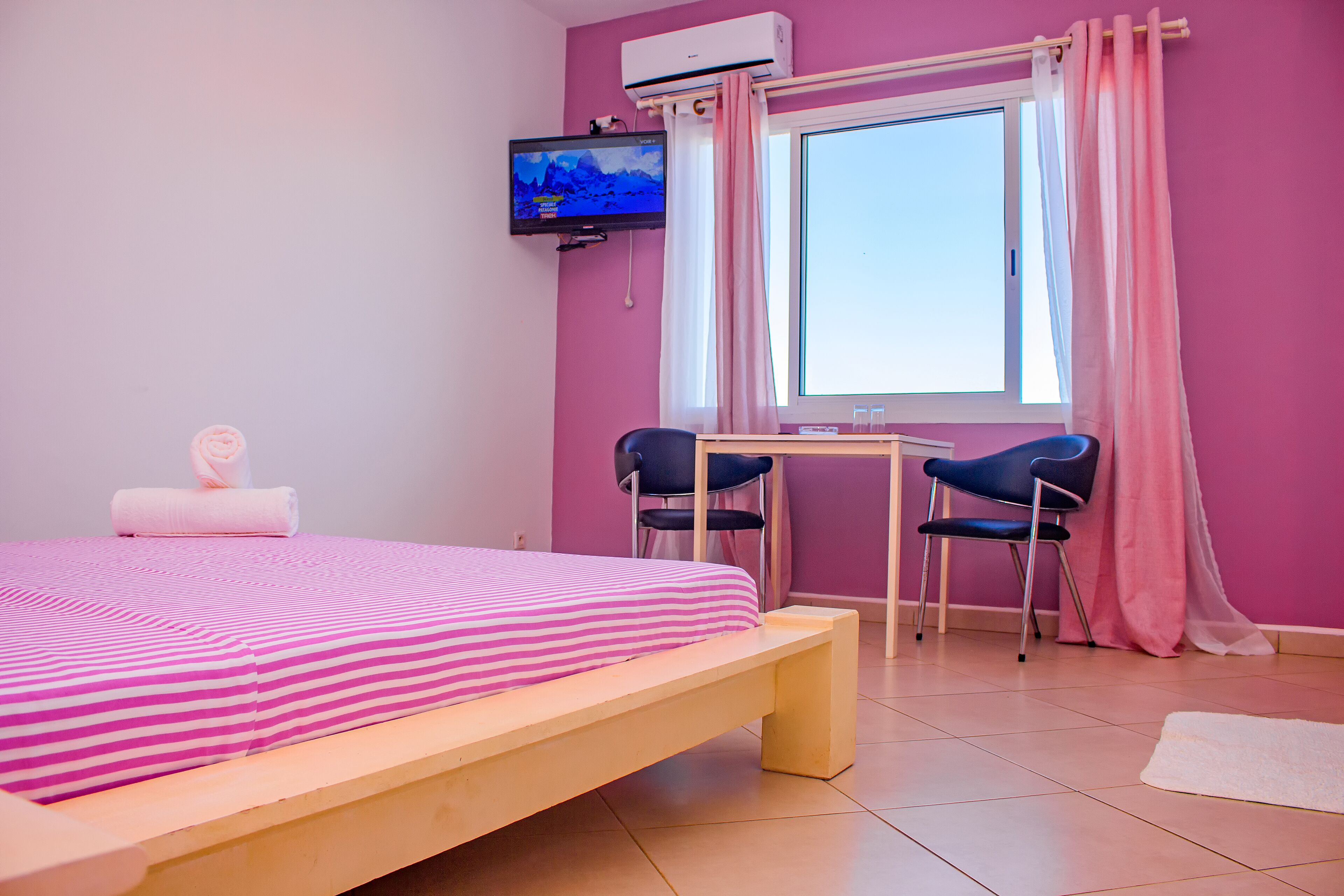 Una habitación con cama, televisor, ventana con cortinas, escritorio y dos sillas.