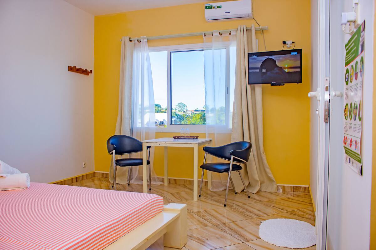 A room with a bed, a table, two chairs, a TV, and a window with curtains.