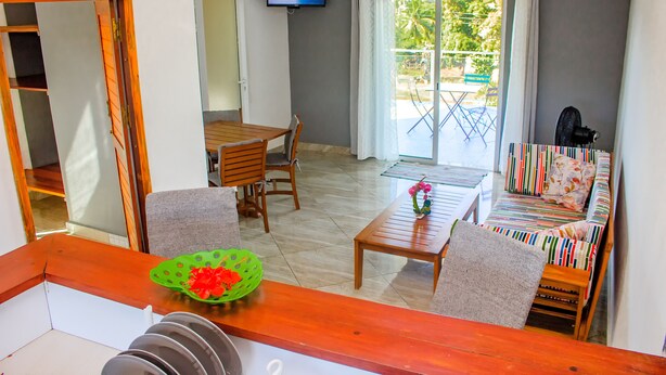 Una habitación con una mesa de comedor de madera, sillas, un sofá, un televisor montado en la pared y vistas al exterior a través de un ventanal.