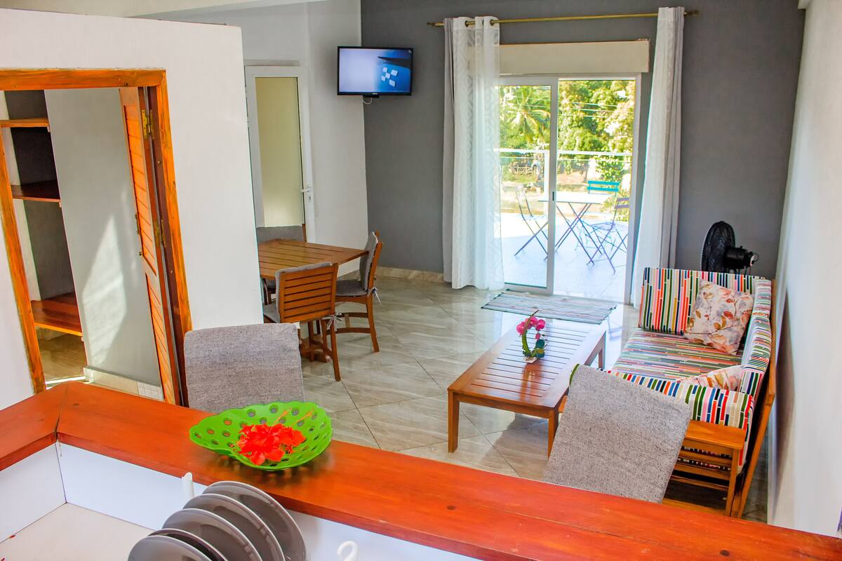 A room with a wooden dining table, chairs, a sofa, a TV mounted on the wall, and a view of the outside through a glass door.