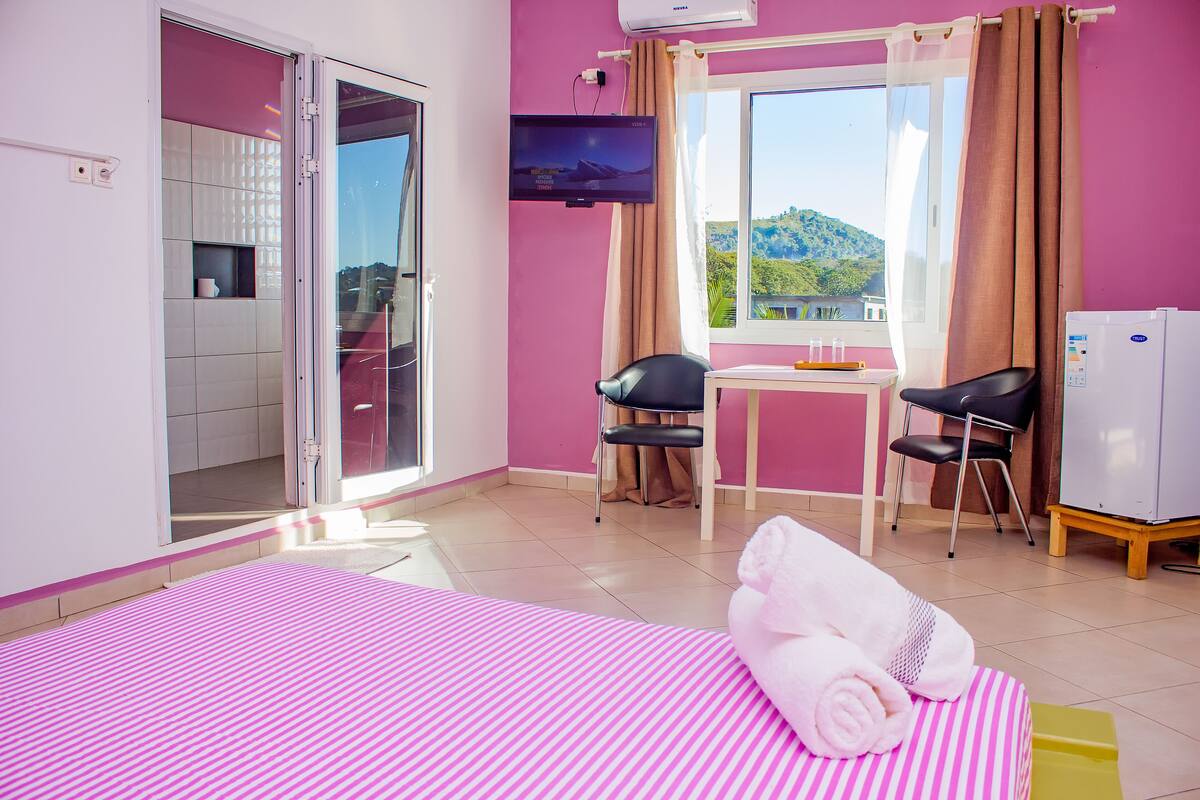 A hotel room with a bed, a TV, a table with chairs, and a view of a mountain.