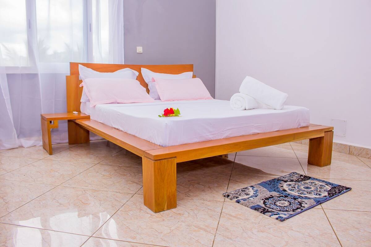 A wooden bed with white bedding and pillows, a bedside table, and a patterned rug on a tiled floor.