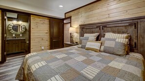 Un dormitorio con cabecera de madera, una cama grande con colcha a cuadros, una mesita de noche con lámpara y un espejo sobre el tocador.